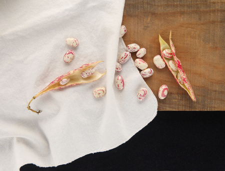Overhead of cranberry beans, some in the shell, others loose on a white cloth on a wood surface with...