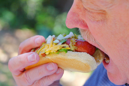 A senior man bites into a hot dog topped with raw onions, relish, lettuce and grated cheeseの写真素材