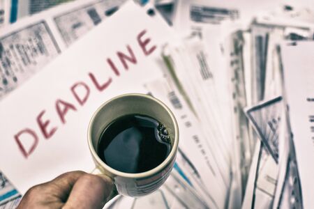 Deadline written on paper and a cup of coffee in hand against a background of scattered papers.  の写真素材
