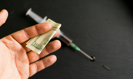 Money for drugs. A man's hand stretches dollars against the background of a syringe with drugs on a dark background.の写真素材