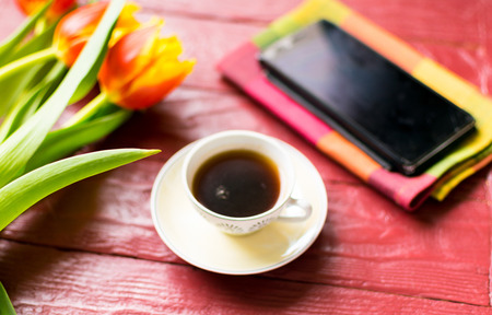 A cup of coffee and tulips and a mobile phone on a burgundy wooden background.の写真素材
