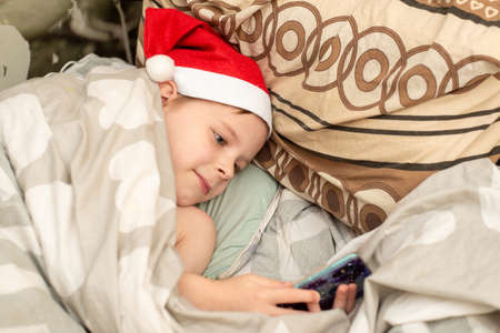 A boy in a Santa hat lies in bed and plays on the phoneの写真素材