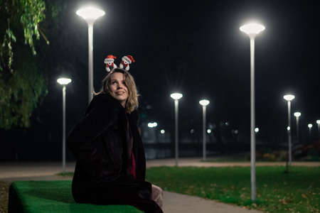 A woman with a New Year's decoration on her head sits at night in a lighted park. The atmosphere of...