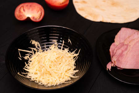 Cooking tortilla with cheese and ham. Pita, grated cheese, ham, tomatoes on a black wooden background.の写真素材