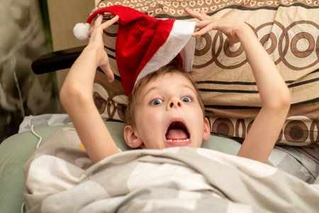 Crazy Christmas. A boy in a Santa hat is lying in bed making faces.の写真素材