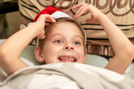 A satisfied, smiling boy lies in bed in a Santa hat. The joyful feeling of the holidays - New Year, Christmas.の写真素材
