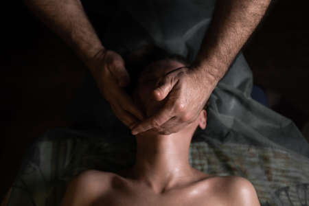 Professional facial massage. Male masseur makes procedures on a female face on a dark backgroundの写真素材