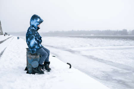 A young boy sits in the winter sadly near the frozen river.の写真素材