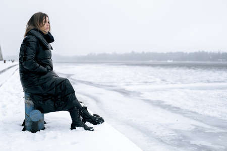 A woman sits alone on the banks of the river in winter. Feminine loneliness, sadness, anticipation,...