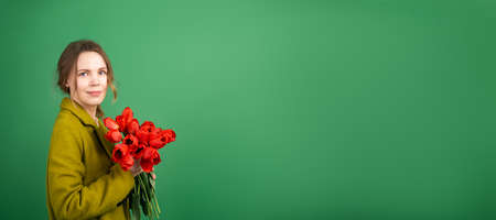 An adult woman stands sideways with a bouquet of tulip flowers on a green background and looks into the camera. Banner. Place for your text. Mother's day, holiday, congratulation, victory day, spring mood conceptの写真素材