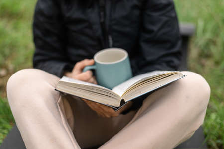 A woman with a cup and a book in the park sits on the green grass on a cool autumn day.の写真素材