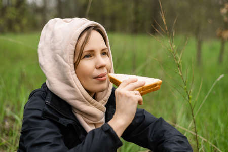 An adult attractive woman eats a sandwich outdoors in the forest, park. Snack on a hike, on a walk.の写真素材