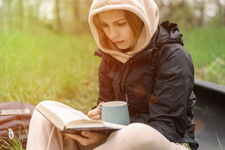 A woman with a cup and a book in the park sits on the green grass on a cool autumn day.