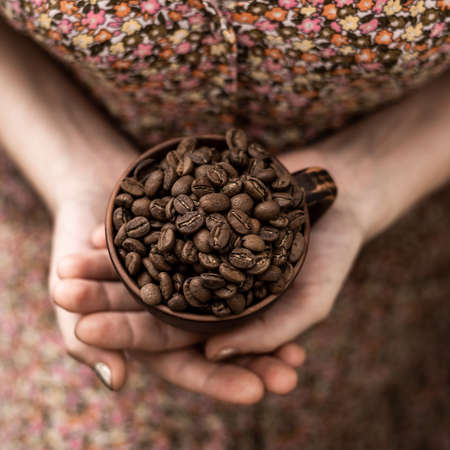 Coffee beans in a cup in female hands on a brown background.の写真素材
