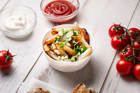 Fried potatoes in a white plate with green onions, ketchup on a white wooden background.