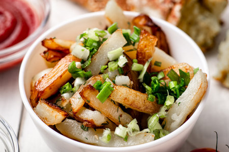 Fried potatoes in a white plate with green onions, parsley, tomatoes on a white wooden background.