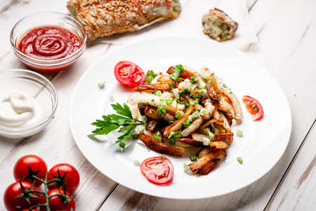 Fried potatoes in a white plate with green onions, parsley, tomatoes on a white wooden background.
