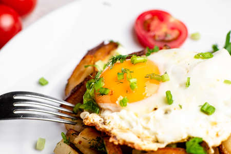 Fried potatoes with egg with liquid flowing yolk, herbs and tomatoes on a white plate.の写真素材