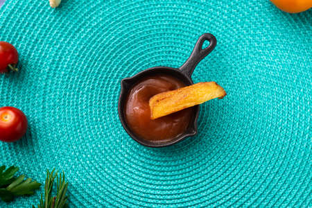 Fried potatoes dipped in ketchup on a turquoise background. There are various vegetables nearby - tomatoes, herbs.の写真素材