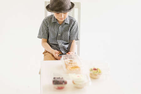 A young boy in a shirt and hat sits in front of cakes with a phone in his hands.の写真素材