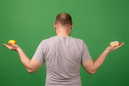 A man holds a cake and an apple on a green background. The choice between healthy and unhealthy food. Healthy lifestyle or obesity.back view.の写真素材