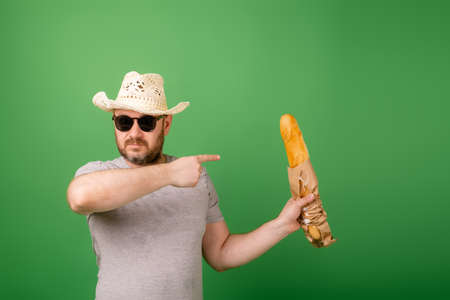 A man in a hat and sunglasses shows a French baguette on a green background.の写真素材