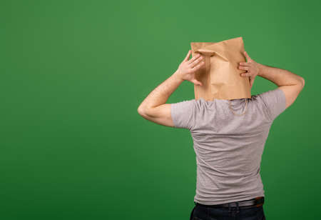 An unrecognizable man with a paper bag on his head grabbed his head against a green background. Place for your text. mental health, greenwashing, disinformation conceptの写真素材