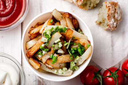 Fried potatoes in a white plate with green onions, ketchup on a white wooden background.