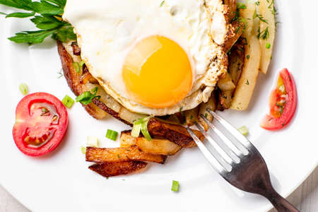Fried potatoes with egg, herbs and tomatoes on a white plate on a white wooden background.