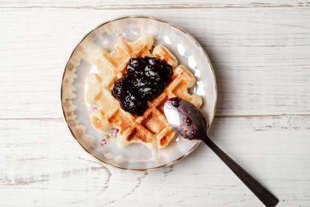 Belgian waffles with blueberry or blackcurrant jam on a plate with a spoon on a white wooden...