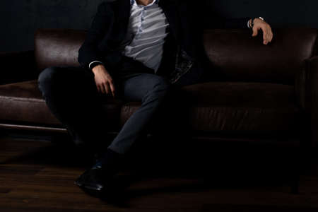 An unrecognizable man in stylish clothes sits relaxed on the sofa in the dark.の写真素材