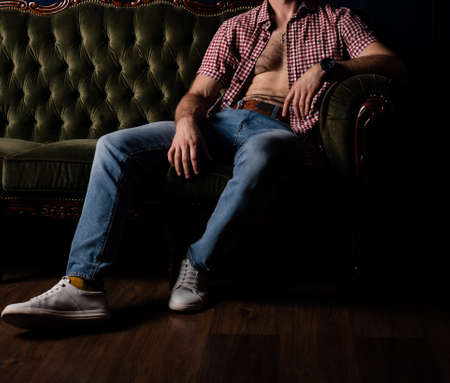 An unrecognizable man with an unbuttoned shirt sits on a sofa in a dark room.の写真素材