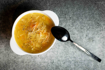 Chicken soup with noodles, potatoes, onions and carrots in a white deep plate on a gray background.