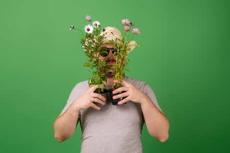 A male gardener, florist in a hat and sunglasses holds fresh flowers in pots on a green background. Plant breeding, sale.の写真素材