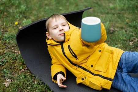 A young boy in a jacket drinks from a cup in the green forest at a picnic.の写真素材