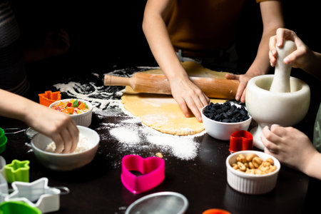 Children cook cookies together. The shortcrust pastry is rolled out with a rolling pin. Various forms for cutting cookies, nuts, raisins, candied fruits on a dark kitchen table.の写真素材