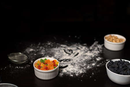 Ingredients for making cookies on a dark background. Flour on the table, candied fruits, raisins,...