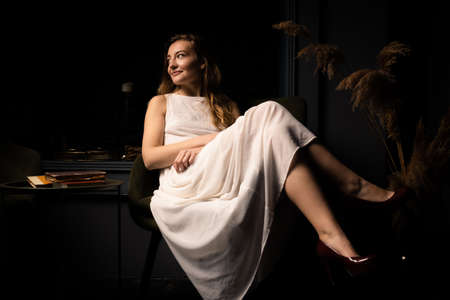 A young woman in a white dress sits relaxed on an armchair in a dark room.の写真素材