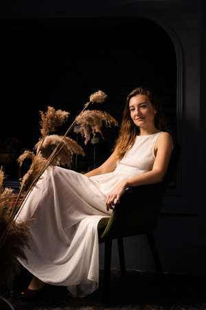 A young woman in a white dress sits thoughtfully in an armchair in a dark room. Solitude, introspection concept.の写真素材