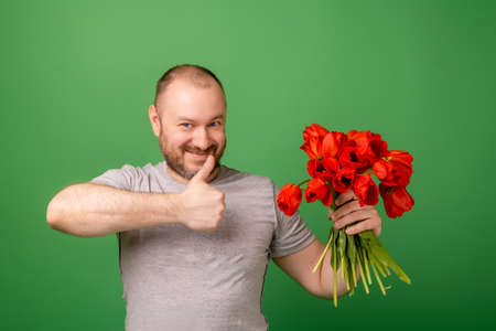 Unshaven happy middle-aged man holding a bouquet of red tulip flowers and an approving gesture with the other hand on a green background. Celebrate, apologize concept.の写真素材