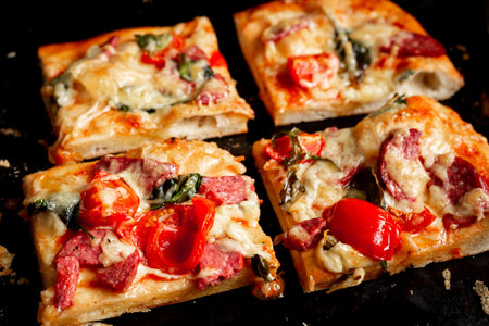 Sliced square pieces of pizza, pie, with tomatoes, sausage, cheese cooked in the oven on a black baking sheet.の写真素材
