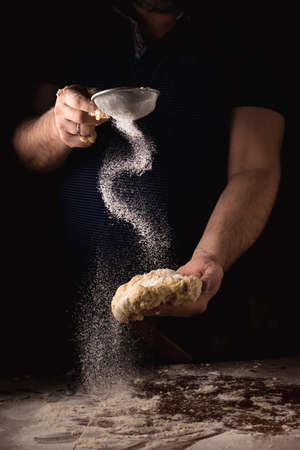 Cooking, kneading dough. Male hands sift flour onto the dough on a dark background.