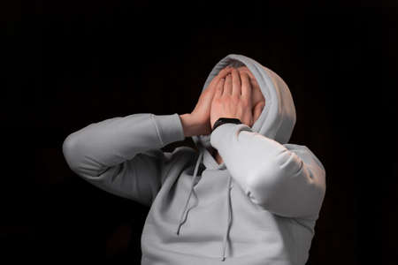 The young man covered his face with his hands. Fear, depression, despair concept. dark background. A man in a sweatshirt with a hood over his head.の写真素材