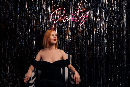 Stylish middle-aged woman sits in a chair. Party inscription on the luminous wall. Bare shoulders. A...