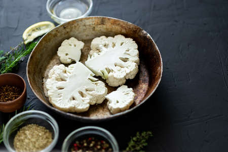 Cauliflower steak cooking. Raw cut cauliflower lie in a frying pan. Olive oil, herbs, various spices nearby. dark background. Place for text.の写真素材