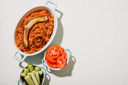 Jollof rice with fried banana. Fresh vegetables - tomato and cucumber. white background. Traditional Nigerian rice food with tomatoes, onions and spices.の写真素材