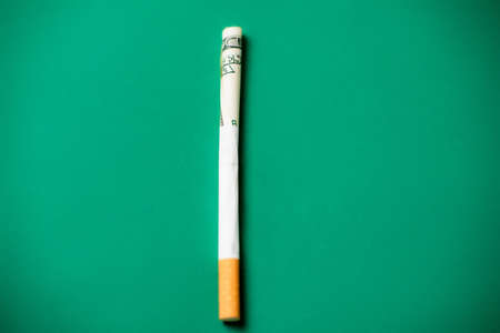 smoking and money. Rolled up dollar bill in a cigarette. green background. The high cost of smoking cigarettes concept.の写真素材