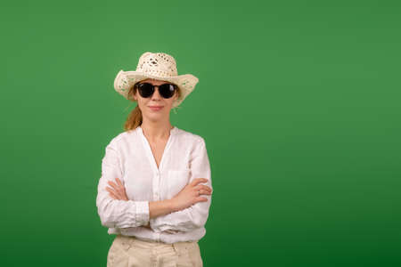 Calm confident middle-aged woman in a cap, white shirt and sunglasses on a green background. Place...