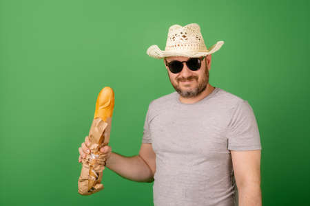 A man in a hat and sunglasses with a baguette in his hands on a green background.