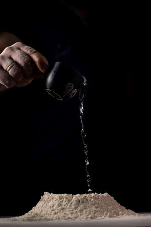 Dough preparation. A man adds water to flour against a dark background.の写真素材